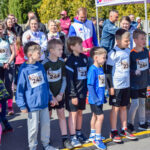 Grupa dzieci w kolorowych strojach stoi na linii startu biegu. W tle widoczny tłum dorosłych i zielone drzewa. Dzień jest słoneczny.