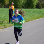 Dziewczynka biegnie w zielono-niebieskiej bluzie z numerem 231. W tle zielone pole i dwóch obserwatorów. Słoneczny dzień.