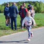 Dziewczynka biegnie w zawodach na asfaltowej drodze, ubrana kolorowo. W tle zielone pole i ludzie obserwujący. Ciepły, słoneczny dzień.