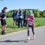 Dziewczynka w różowej bluzie z numerem startowym biegnie przy drodze. W tle zielone drzewa i grupa osób. Słońce jasno świeci.