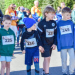 Grupa dzieci w kolorowych strojach sportowych z numerami startowymi stoi na drodze. Za nimi widoczny tłum dorosłych. Słoneczna pogoda, zieleń w tle.