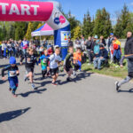 Dzieci startują w biegu pod różowym łukiem z napisem START. Tło to widownia i zielone drzewa. Dzień jest słoneczny.