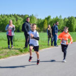 Dwóch chłopców biegnie w zawodach na wiejskiej drodze. Jeden ma biały strój, drugi pomarańczową bluzę. W tle zielone drzewa i kibice.
