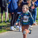 Na zdjęciu uśmiechnięty chłopiec biegnie, wokół dopingujący ludzie. Tło to zielona trawa. Dominują kolory niebieski i zielony.