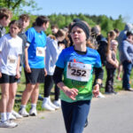 Młoda osoba w niebieskiej koszulce z numerem 252 biegnie w wyścigu. W tle widać zielone drzewa i kibiców w różnych ubraniach. Słoneczna pogoda podkreśla atmosferę sportowego wydarzenia na świeżym powietrzu.