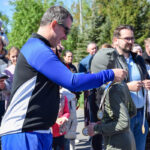 Grupa ludzi na świeżym powietrzu, mężczyzna w niebieskiej bluzie wręcza medale dzieciom. W tle zieleń i drzewa. Słoneczny dzień.