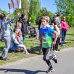 Biegacz w niebiesko-zielonej koszulce na trasie biegu w parku, wokół widzowie. Zielone drzewa w tle, słoneczna pogoda.