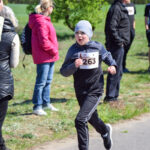 Chłopiec w czapce biegnie podczas wyścigu na świeżym powietrzu. Nosi czarny strój, numer startowy 263. W tle trawa i stojący ludzie.