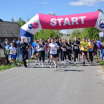 Na zdjęciu grupa biegaczy startuje spod różowo-białego łuku startowego. Uczestnicy biegną po asfalcie, otoczeni kibicami. W tle widać drzewa i fragmenty budynków.