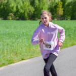 Dziewczyna w różowej bluzie biegnie na asfaltowej drodze. Zielone pola i drzewa w tle. Na koszulce numer 173.