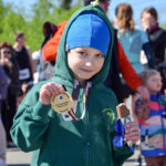 Chłopiec w zielonej bluzie i niebieskiej czapce trzyma medal oraz baton. W tle rozmyty tłum i zielone drzewa. Słoneczny dzień.
