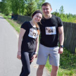 Para w sportowych strojach z numerami startowymi pozuje na asfaltowej drodze. Tło stanowią zielone drzewa i drewniany płot. Jasne niebo podkreśla słoneczną pogodę.