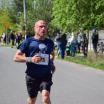 Biegacz w niebieskiej koszulce z numerem startowym na asfaltowej drodze. W tle drzewa i widzowie. Słoneczny dzień.