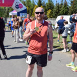 Mężczyzna w czerwonej koszulce, z medalem, pozuje na zawodach biegowych. W tle widać ludzi i zielone drzewa. Niebieskie niebo i różowy łuk startu.