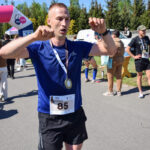 Biegacz w niebieskiej koszulce z medalem na piersi, w tle zielone drzewa i niebo. Na asfalcie droga, wokół uczestnicy biegu.