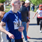 Mężczyzna w niebieskiej koszulce z numerem startowym trzyma butelkę wody. W tle biegacze i zielone drzewa. Słoneczny dzień.