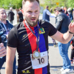 Mężczyzna w sportowej koszulce z medalem na tle uczestników wydarzenia. Ubrany w czarno-czerwono-niebieską koszulkę. W słoneczny dzień na świeżym powietrzu.