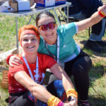 Dwie uśmiechnięte kobiety siedzą w trawie, ubrani w sportowe stroje. Jedna ma czerwoną koszulkę, druga niebieską. W tle metalowe ogrodzenie i pudełka.