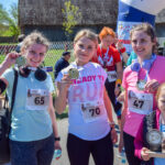 Trzy kobiety z medalami po biegu, w tle zielone drzewa i budynek. Mają stroje sportowe w jasnych kolorach. Uśmiechnięte, podniosły puchary.