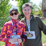 Zdjęcie przedstawia dwie uśmiechnięte osoby w sportowych strojach. Kobieta w kolorowej kurtce trzyma butelkę wody. W tle siatka i zielone drzewa.