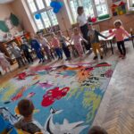 Dzieci stoją w kole na kolorowym dywanie w przedszkolu. W tle widać okna i dekoracje na ścianach. Pomieszczenie jest jasne i przestronne.