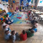 Grupa dzieci siedzi w kole na kolorowym dywanie w przedszkolu. W tle regały z zabawkami i książkami. Ściany ozdobione dekoracjami.