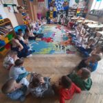 Grupa dzieci siedzi w kręgu na kolorowym dywanie w przedszkolu. W tle zabawek, stoły i dekoracje. Ściany ozdobione plakatami i balonami.