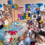 Sala przedszkolna z kolorowym dywanem. Dzieci siedzą przed nauczycielką w kwiecistym stroju. Kolorowe ozdoby i zabawki na półkach.