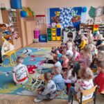 Na zdjęciu grupa dzieci w przedszkolu słucha opowieści. Kolorowy dywan z motywem zwierząt i regały z zabawkami w tle. Nauczycielka po lewej w kapeluszu przy tablicy.