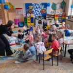 Grupa dzieci siedzi na kolorowym dywanie, uważnie patrząc na kobietę. W tle regały z zabawkami i kolorowe pudełka. Sala ozdobiona jest niebieskimi balonami i rysunkami.