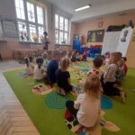 Grupa dzieci siedzi na kolorowym dywanie w przedszkolnej sali. Jasne ściany i duże okna wpuszczają dużo światła. Nauczycielka stoi z przodu pokoju, dekoracje w tle.