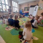 Dzieci siedzą na kolorowym dywanie w przedszkolu. Tło stanowi jasna ściana z oknami. Nauczycielka stoi z przodu.