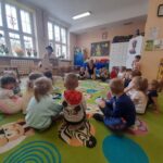 Sala przedszkolna z dziećmi siedzącymi na kolorowym dywanie. Na tle jasnych ścian i dużych okien są szafki i zabawki. Nauczyciel prezentuje coś na stoliku.