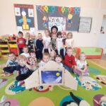 Grupa uśmiechniętych dzieci siedzi na kolorowym dywanie z postaciami. W tle szafki i ozdoby ścienne. Dzieci pozują przy drewnianej książce.