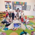 Grupa dzieci na kolorowym dywanie w przedszkolu, w tle jasna ściana z dekoracjami i półki z zabawkami.