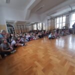 Dzieci siedzą na drewnianej podłodze w jasnej sali gimnastycznej. Na lewo starsza kobieta z aparat. Ściany mają drabinki gimnastyczne.