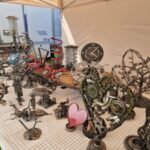 3B7qaJpY Stoisko z metalowymi rzeźbami na białym obrusie, w tym zegary i serca. Tło beżowe, namiot. Kolory srebrny, czerwony i zielony.