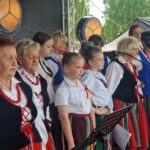 500281758_728062479569276_2748257806727929296_n Grupa osób w tradycyjnych strojach ludowych śpiewa na scenie. Dominują kolory biały, czerwony i czarny. W tle widać zielone drzewa.