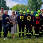 500302924_728976972811160_7929166415505538522_n Grupa osób, w tym dzieci w strojach strażackich z zielonymi kaskami, stoi na trawie. W tle namioty i drzewa. 明るい昼間のシーン。