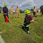 500313062_728980119477512_6734918746338608103_n Grupa osób w strażackich uniformach bierze udział w ćwiczeniach na zielonym trawniku. Dziecko w jasnym hełmie trzyma wąż strażacki. Obserwują ich inni w beżowych mundurach na tle drzew i chmur.
