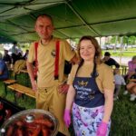 500313557_728990276143163_8442413694818749652_n Dwóch dorosłych w jasnych ubraniach stoi pod zielonym namiotem. Na grillu pieką się kiełbaski. W tle widać siedzących ludzi i zieleń.