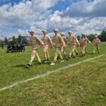 500331366_728988749476649_8912582835774581282_n Grupa ludzi w beżowych mundurach maszeruje po zielonym trawniku. W tle siedząca grupa w czarnych strojach. Niebo jest błękitne z białymi chmurami.