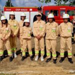 500360638_728977562811101_5049300068166624910_n Grupa młodych strażaków w beżowych mundurach z białymi kaskami stoi przed czerwonym wozem strażackim. Za nimi niebo z chmurami. Na mundurach zielone napisy STRAŻ.