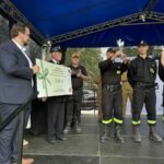 500377177_728972976144893_8974648647725209188_n Na scenie grupa strażaków odbiera nagrodę. Mężczyzna trzyma duży czek na 1500 zł. Tło w kolorze niebieskim i czarnym.