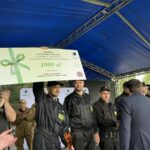 500378900_728972662811591_1663411518306775300_n Nie wiem, kto jest na zdjęciu, ale widzę grupę ludzi w ciemnych uniformach stojących na scenie z niebieskim zadaszeniem. Jeden z nich trzyma dużą nagrodę pieniężną o wartości 1000 zł z zieloną kokardą. W tle znajdują się drzewa