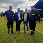 500392578_728974132811444_3093390483510477236_n Grupa czterech mężczyzn stoi na zielonej trawie. Dwóch nosi czarne mundury strażackie. W tle widać scenę i drzewa pod pochmurnym niebem.