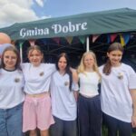 500472749_728062209569303_3153220673484540595_n Grupa pięciu młodych osób w białych koszulkach stoi przed zielonym namiotem z napisem Gmina Dobre. Tło zdobią kolorowe balony i girlandy. Namiot jest ustawiony na trawie podczas wydarzenia na świeżym powietrzu.