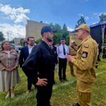 500473212_728990766143114_7626066268972505526_n Grupa osób na otwartym terenie rozmawia w słoneczny dzień. Mężczyzna w beżowym mundurze stoi naprzeciw osoby w czerni. W tle są zielone drzewa i niebieskie niebo.