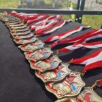 500488947_727959632912894_5499558117951599256_n Na czarnym tle leży rząd złotych medali z biało-czerwonymi wstążkami. Medale mają czerwono-niebieskie detale i wzór nawiązujący do strażaków. W tle widać zieloną trawę przez siatkę.