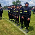 500502241_728990549476469_1158872987244764703_n Strażacy w czarnych mundurach stoją w szeregu na zielonym trawiastym boisku. Na mundurach żółte napisy STRAŻ. W tle niebo z chmurami i kilka budynków.